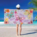 A person at the beach holds a towel decorated with colorful butterflies and flowers, featuring the name \“Sylvia\“ on it.