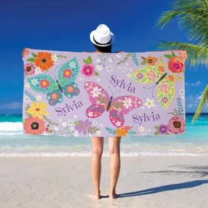 A person at the beach holds a towel decorated with colorful butterflies and flowers, featuring the name \“Sylvia\“ on it.