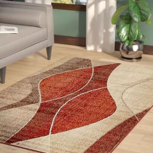 A 5' x 8' geometric area rug with curved patterns in shades of red, beige, and brown on a hardwood floor near a gray couch and a potted plant.