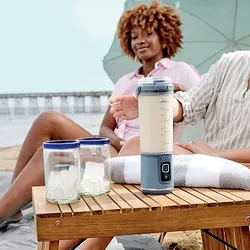 A portable blender with a digital display is on a wooden table next to glass jars of ice, set outdoors on the beach with two people relaxing in the background.