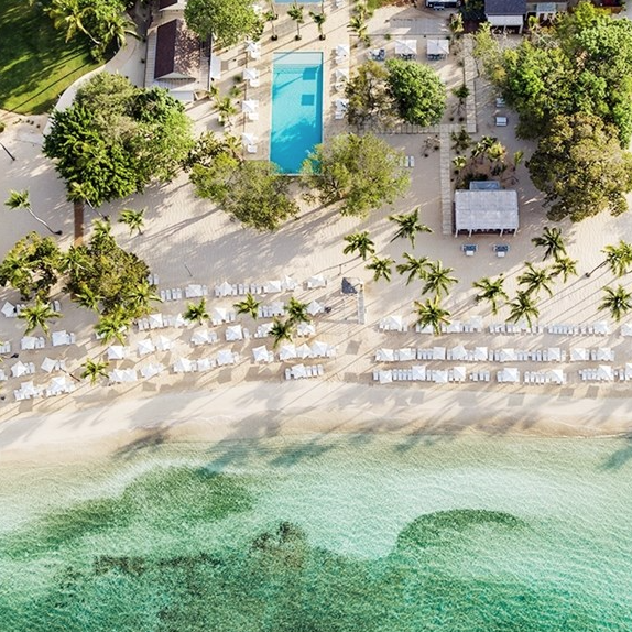 Aerial view of a tropical beach resort featuring a long swimming pool, rows of sun loungers with umbrellas, palm trees, and turquoise water.