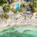 Aerial view of a tropical beach resort featuring a long swimming pool, rows of sun loungers with umbrellas, palm trees, and turquoise water.