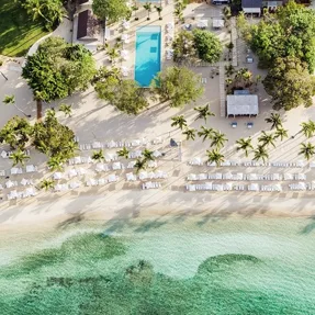 Aerial view of a tropical beach resort featuring a long swimming pool, rows of sun loungers with umbrellas, palm trees, and turquoise water.