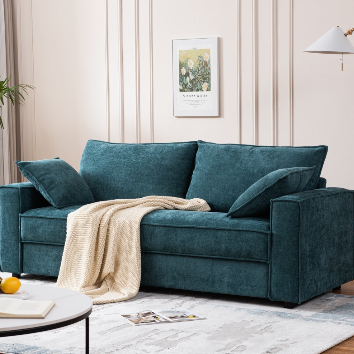 A teal fabric sofa with matching cushions is adorned with a cream-colored knitted throw. It is set in a stylishly decorated room with a white circular coffee table and wall art.