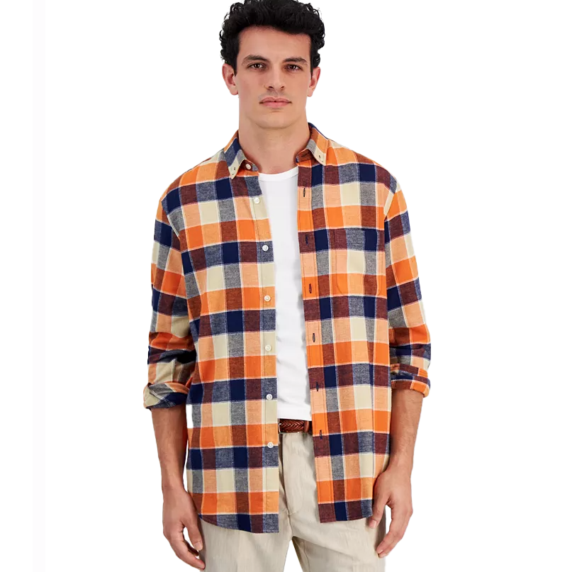 A man is wearing a long-sleeve, button-up flannel shirt with an orange, navy, and cream plaid pattern, layered over a white T-shirt and paired with beige pants.