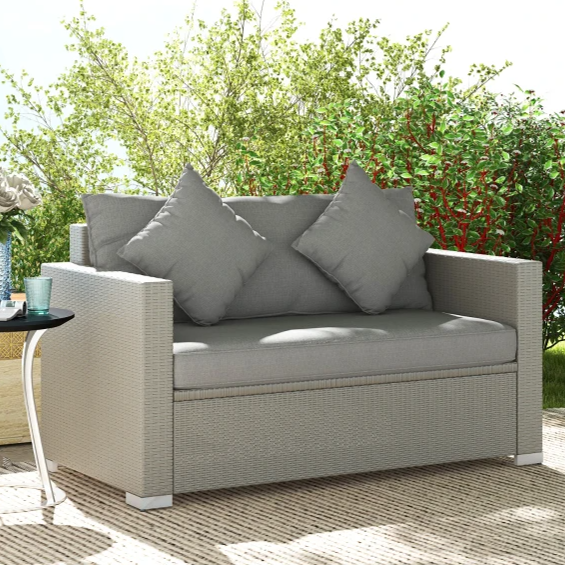A light gray two-seater outdoor sofa with two matching cushions is placed on a patio, accompanied by a small round side table with roses in a vase and cups, partially shaded by tree branches.