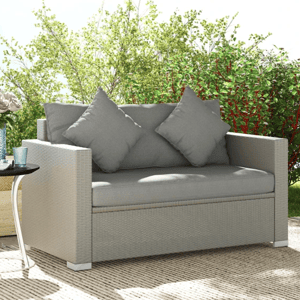 A light gray two-seater outdoor sofa with two matching cushions is placed on a patio, accompanied by a small round side table with roses in a vase and cups, partially shaded by tree branches.