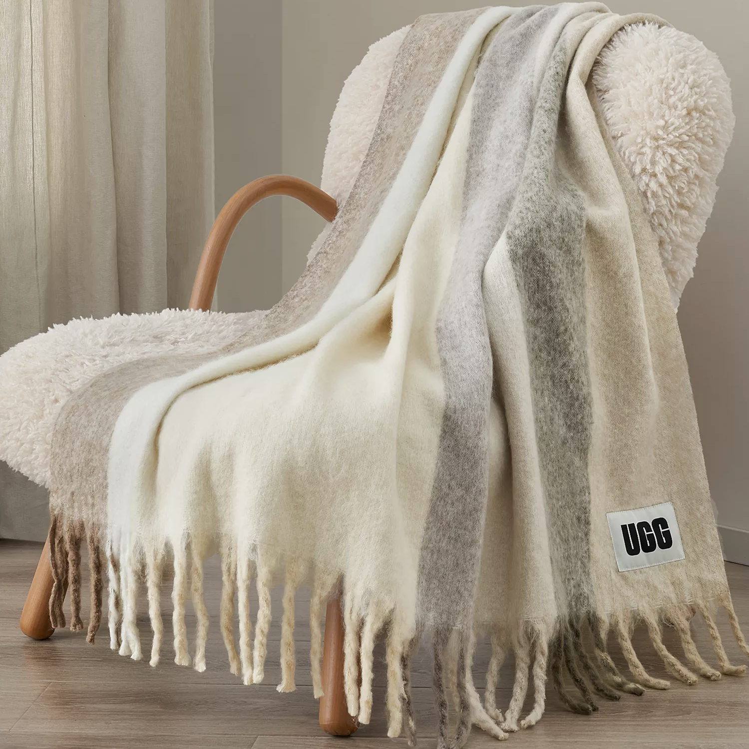 A striped throw blanket with fringe detailing is draped over a textured chair. A visible \“UGG\“ label is attached to the blanket.