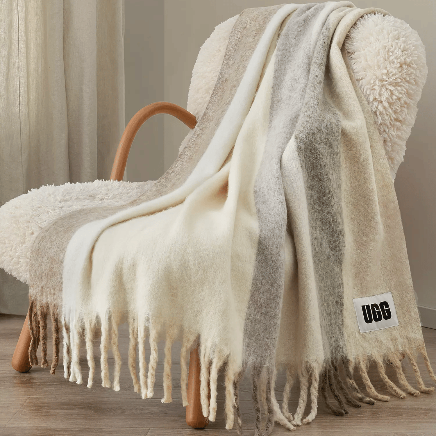 A striped throw blanket with fringe detailing is draped over a textured chair. A visible \“UGG\“ label is attached to the blanket.
