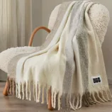 A striped throw blanket with fringe detailing is draped over a textured chair. A visible \“UGG\“ label is attached to the blanket.