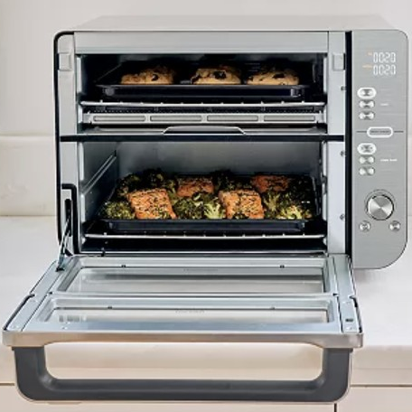 A countertop oven with two cooking racks contains baked goods on the top and a dish with salmon and broccoli on the lower rack.