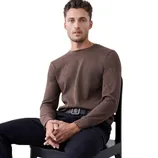 A man sits on a chair wearing a brown long-sleeve sweater and navy pants with a belt, against a plain background.
