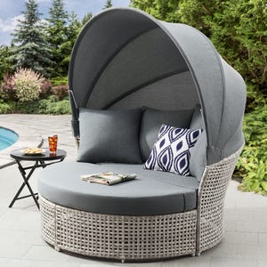Outdoor wicker daybed with canopy and cushions beside a round side table with a beverage and snack.