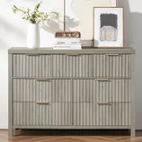 A gray dresser with ribbed drawer fronts is topped with decorative items: a white vase with flowers, a vintage-style radio, and framed artwork.