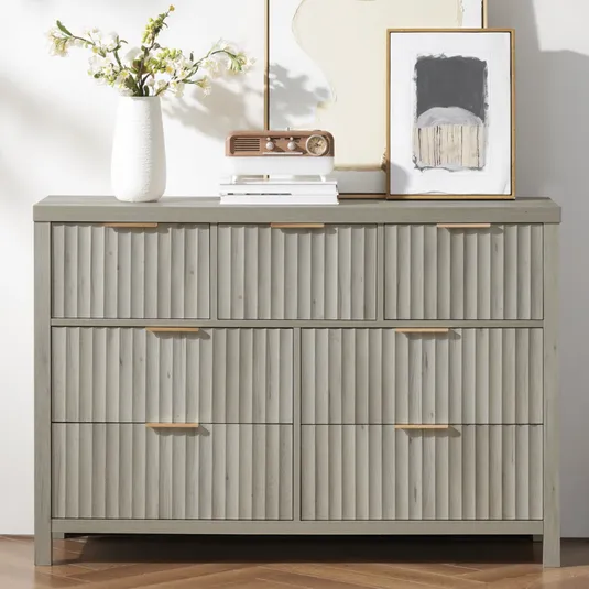 A gray dresser with ribbed drawer fronts is topped with decorative items: a white vase with flowers, a vintage-style radio, and framed artwork.