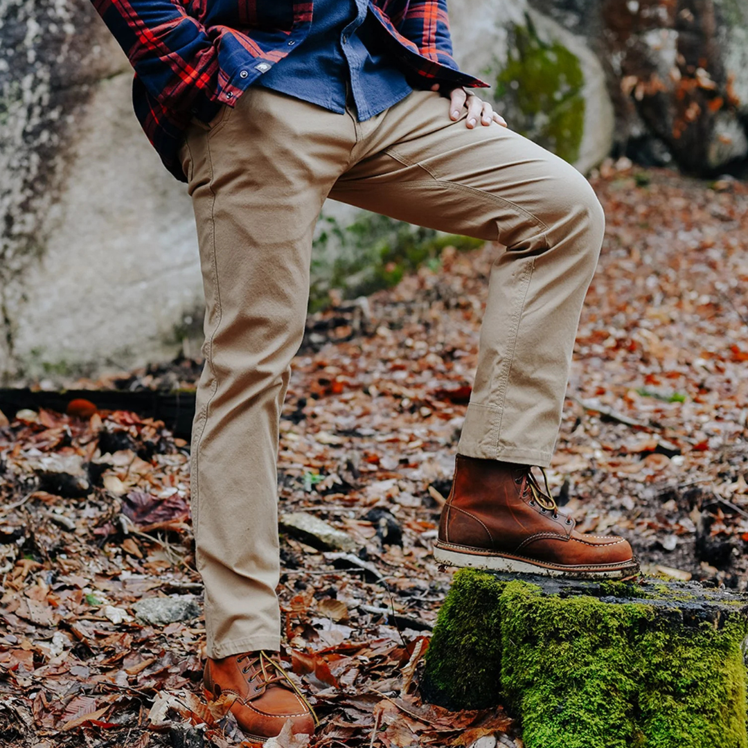 A person is wearing khaki pants, brown leather boots, a blue shirt, and a red plaid jacket, standing on moss-covered ground surrounded by autumn leaves.