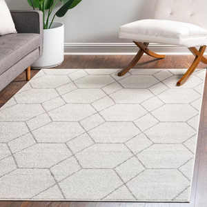 A geometric-patterned area rug on a hardwood floor is placed between a grey sofa and a white, cushioned chair with curved wooden legs, with a tall plant in the background.