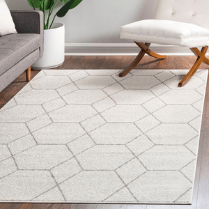 A geometric-patterned area rug on a hardwood floor is placed between a grey sofa and a white, cushioned chair with curved wooden legs, with a tall plant in the background.