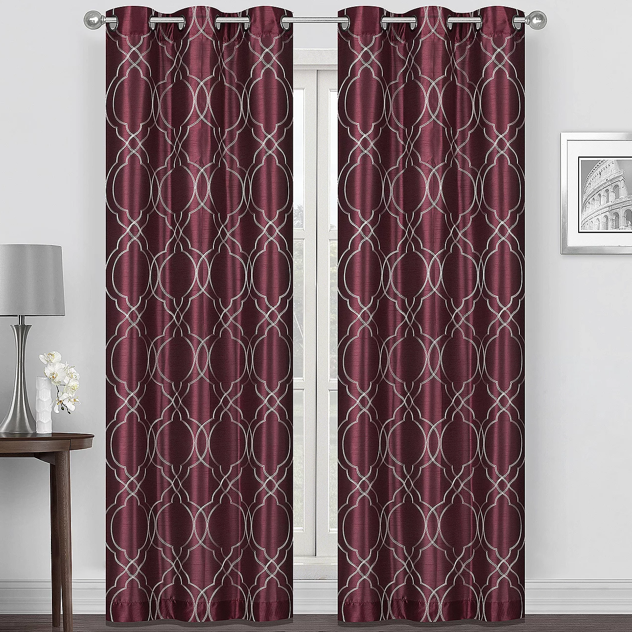 Burgundy curtain panels with a geometric silver pattern, hanging on a rod. They complement a minimalist room featuring a small table, a lamp, and a decorative flower vase.