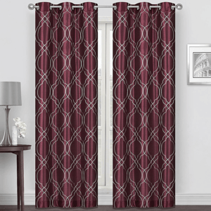 Burgundy curtain panels with a geometric silver pattern, hanging on a rod. They complement a minimalist room featuring a small table, a lamp, and a decorative flower vase.