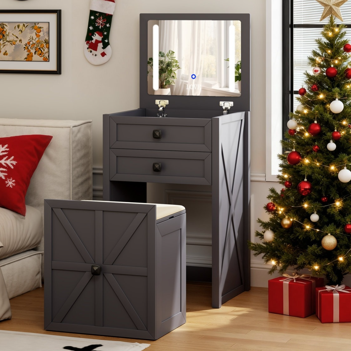 Dark gray vanity set with a foldable mirror and two drawers, accompanied by a matching cushioned stool. Decorated space with a Christmas tree, red and white ornaments, and gift boxes nearby.