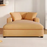 A large, mustard-colored chaise lounge with two matching pillows is placed in a bright living room with a white carpet and decorative wall elements.
