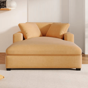 A large, mustard-colored chaise lounge with two matching pillows is placed in a bright living room with a white carpet and decorative wall elements.