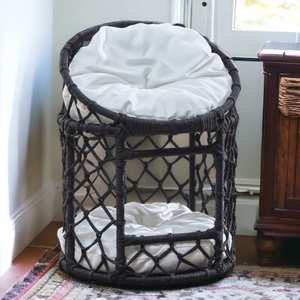 A round, dual-level wicker pet chair with a black woven frame and white cushions is positioned by a window and wooden furniture, resting on a patterned rug.