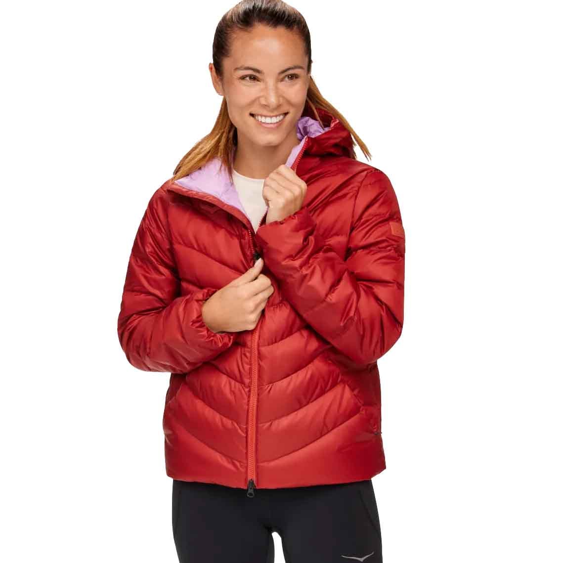 The Hoka Women's Outseeker Down Jacket is a red, puffy winter coat featuring a zip closure and a contrasting inner lining in a lighter shade, designed to provide warmth and comfort.