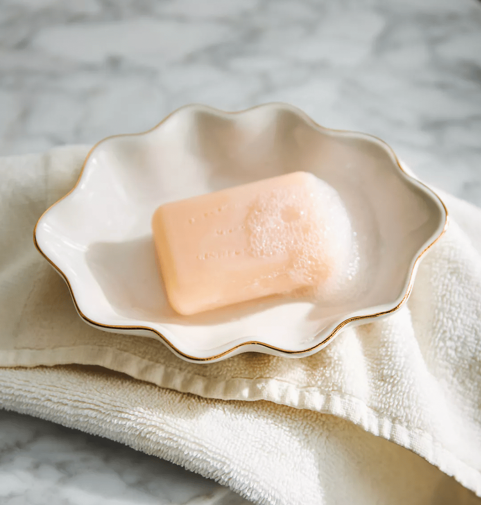 A peach-colored bar of soap with some foam is placed in an elegant, scalloped ceramic soap dish with gold trim, resting on a folded cream towel on a marble surface.