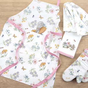 A floral-patterned kitchen set includes an apron, dish towel, oven mitt, and pot holder, all accented with pink trim. A whisk and pastry brush are tucked in the apron pocket.