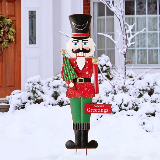 A 36-inch metal nutcracker yard stake, featuring a traditional soldier design with a red and green uniform, black boots, and a tall black hat, holding a small decorated tree and a \“Season's Greetings\“ sign, set against a snowy backdrop.