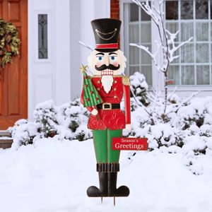 A 36-inch metal nutcracker yard stake, featuring a traditional soldier design with a red and green uniform, black boots, and a tall black hat, holding a small decorated tree and a \“Season's Greetings\“ sign, set against a snowy backdrop.