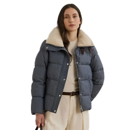 A woman is wearing a blue puffer jacket with a beige sherpa collar and sections. Underneath, she has a light-colored outfit with a belt. She holds a dark bag.