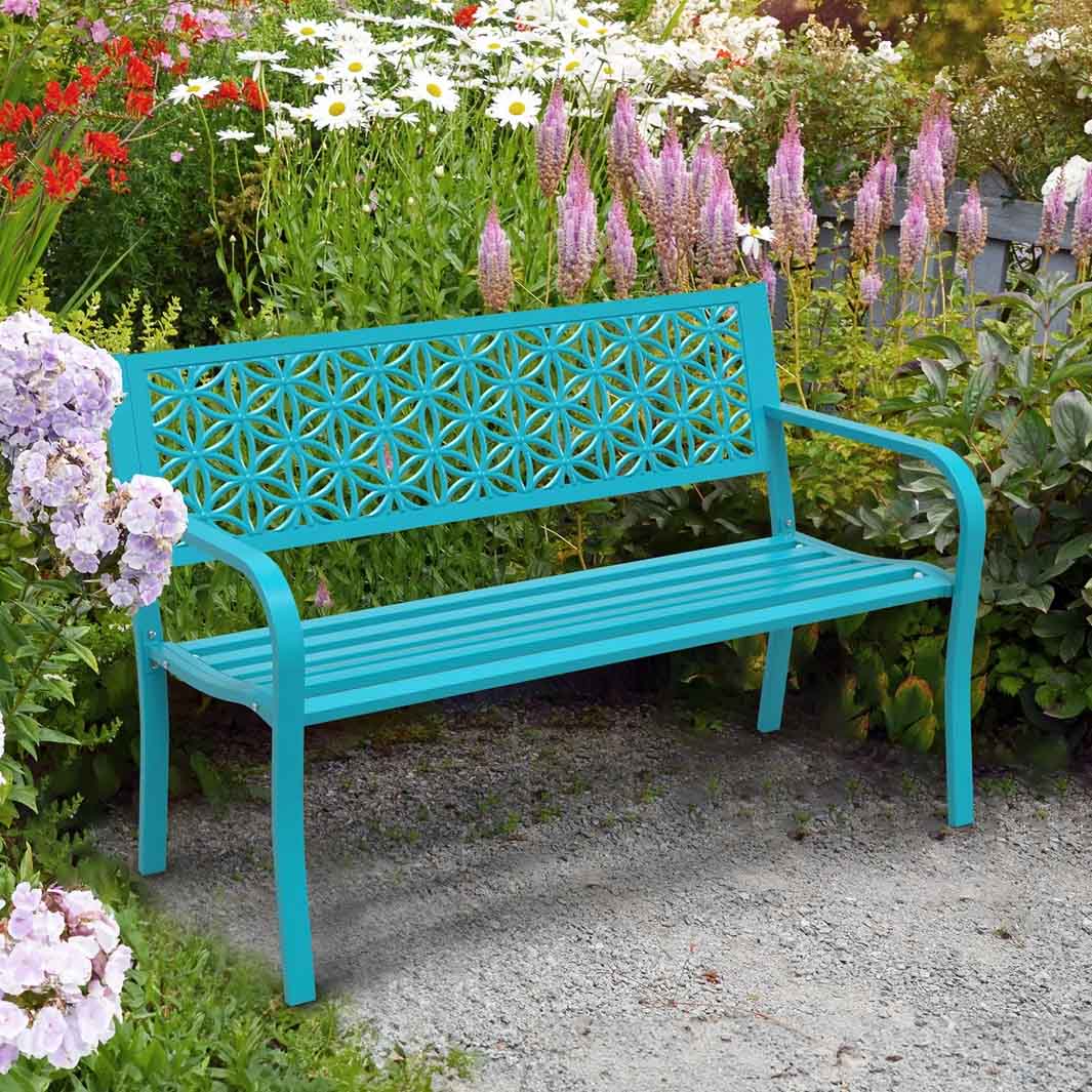 A turquoise metal garden bench features a decorative floral patterned backrest, surrounded by colorful flowers and greenery on a gravel path.