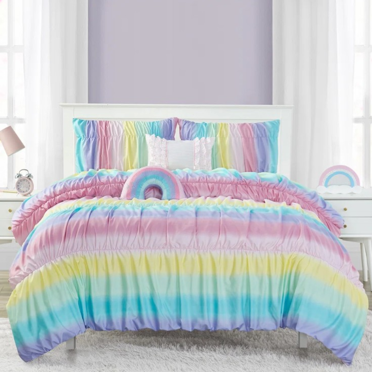 Colorful rainbow-themed bedspread set with ruched texture, featuring matching pillows. Flanked by white nightstands, one with a lamp and clock, set against lavender walls and large windows with sheer curtains.