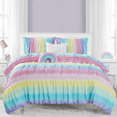 Colorful rainbow-themed bedspread set with ruched texture, featuring matching pillows. Flanked by white nightstands, one with a lamp and clock, set against lavender walls and large windows with sheer curtains.