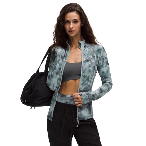 A woman wearing a tie-dye zip-up jacket and black pants holds a black gym bag. She's also wearing a grey sports bra underneath the jacket.