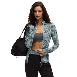 A woman wearing a tie-dye zip-up jacket and black pants holds a black gym bag. She's also wearing a grey sports bra underneath the jacket.
