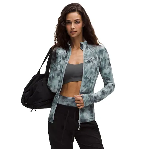 A woman wearing a tie-dye zip-up jacket and black pants holds a black gym bag. She's also wearing a grey sports bra underneath the jacket.
