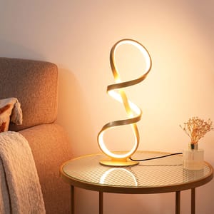 A modern, abstract-shaped LED lamp emits a warm glow on a glass-top side table beside a bed. The table also holds a small vase with dried flowers.