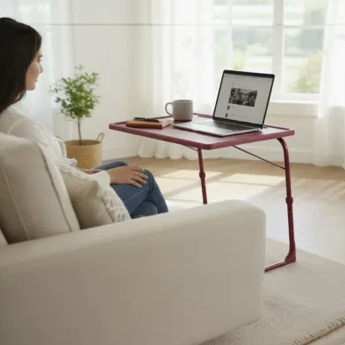 A woman sits on a couch using a red portable table for her laptop, with a mug and phone also on the table. The room features large windows, potted plants, and minimalist decor.
