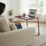 A woman sits on a couch using a red portable table for her laptop, with a mug and phone also on the table. The room features large windows, potted plants, and minimalist decor.