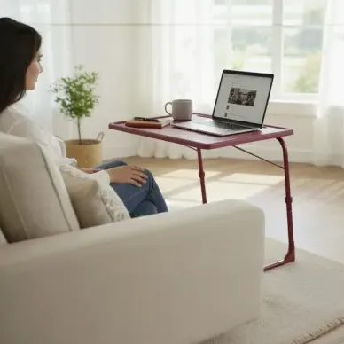 A woman sits on a couch using a red portable table for her laptop, with a mug and phone also on the table. The room features large windows, potted plants, and minimalist decor.