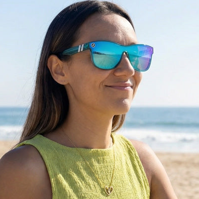 Woman wearing blue reflective sunglasses, with a heart-shaped pendant necklace, standing on a beach.