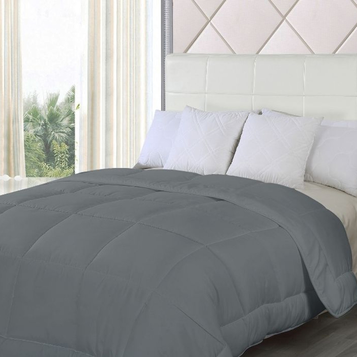 A grey down-alternative comforter is laid on a bed, with white pillows against a white tufted headboard, in a room with a view of trees through a window.