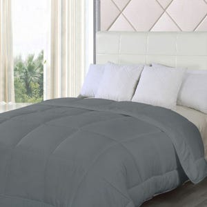 A grey down-alternative comforter is laid on a bed, with white pillows against a white tufted headboard, in a room with a view of trees through a window.