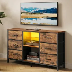 A woman holds a cup on a sofa next to a SONGMICS TV stand, designed to fit TVs up to 55 inches. The stand features drawers, a shelf with a yellow light, and various items like a record player and books.