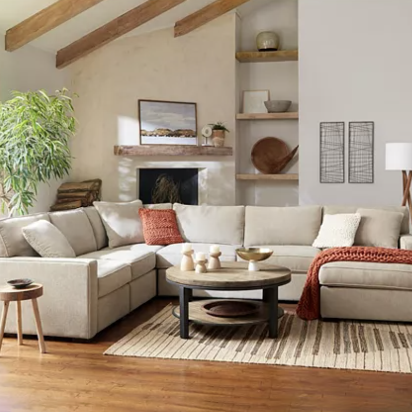 A cozy living room setup with a beige sectional sofa, accentuated by orange and white cushions, a round wooden coffee table on a striped rug, and a tall indoor plant. The space includes a fireplace and wooden shelves with decorative items.