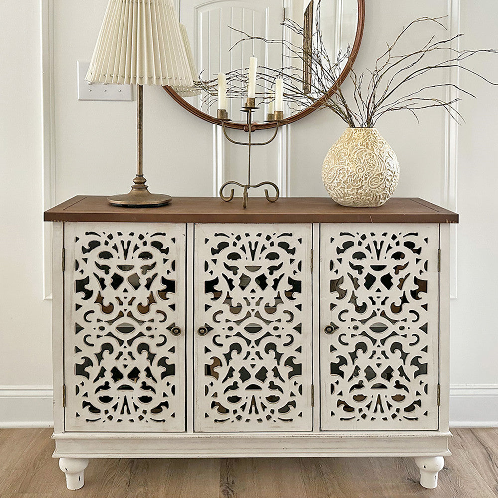 An intricately carved white sideboard with a wooden top is adorned with a pleated lampshade, a branch-filled ornate vase, and a candelabra, set against a round wall mirror.